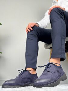 Model u sedećem položaju nosi muške cipele teget C8557-09 uz teget pantalone i belu košulju